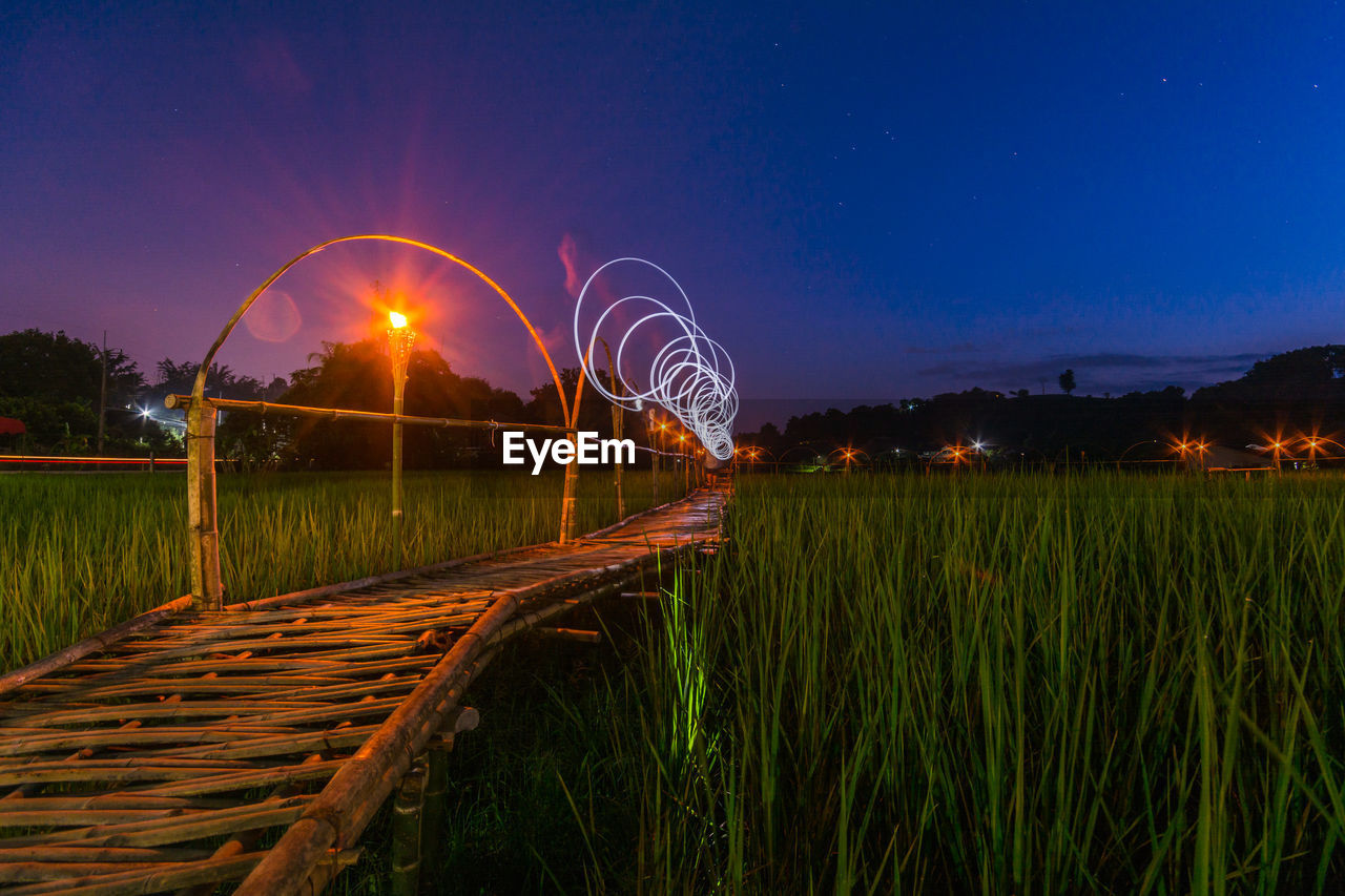 Boardwalk amidst rice field with light painting against sky at dusk