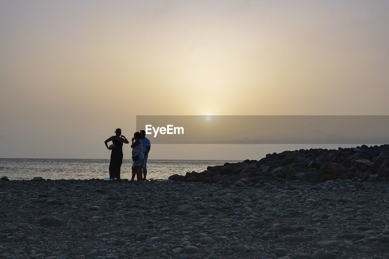 MEN STANDING ON SHORE AGAINST SKY DURING SUNSET