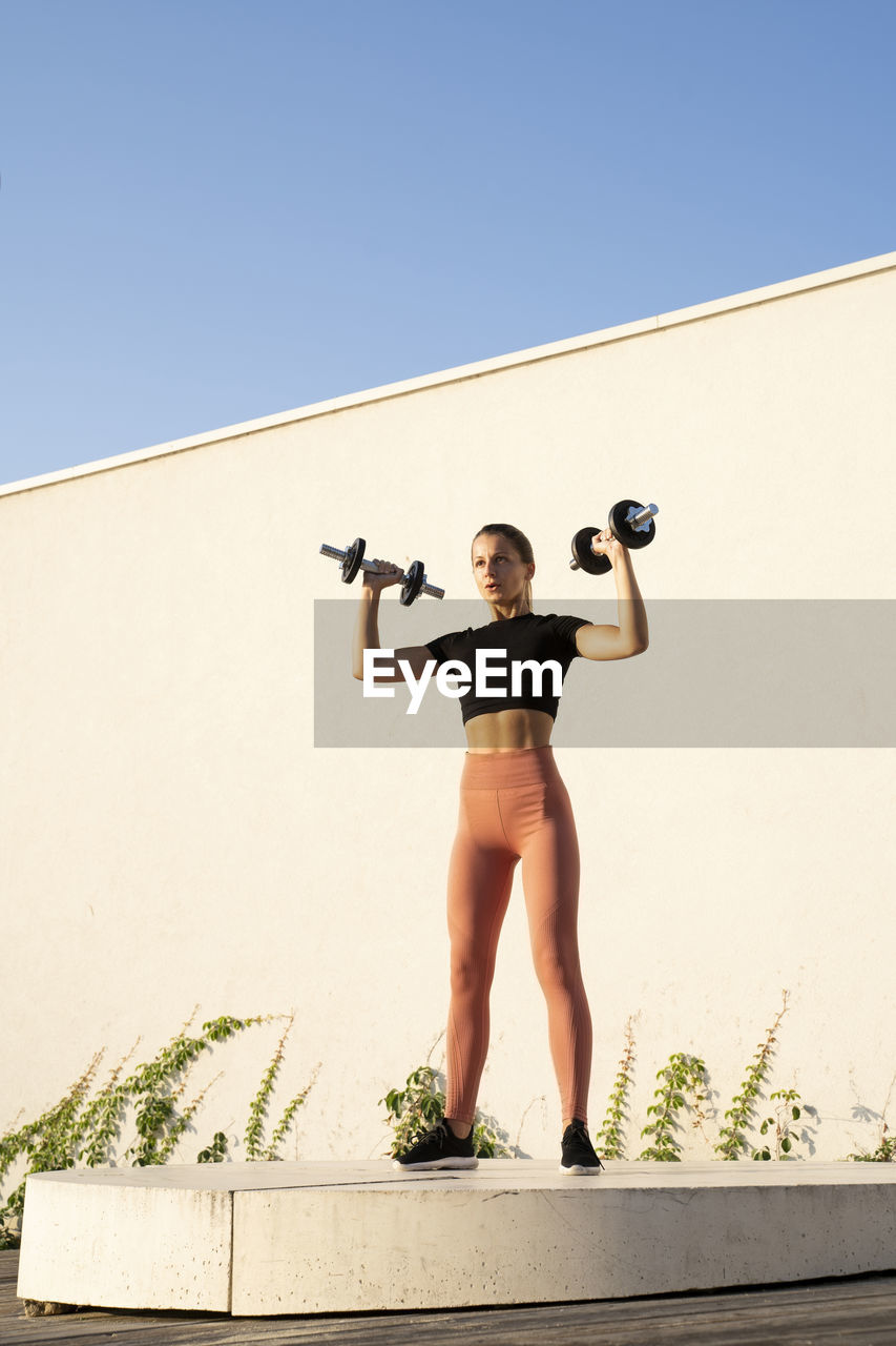 Sportswoman flexing muscle with dumbbell standing on pedestal against wall during sunrise