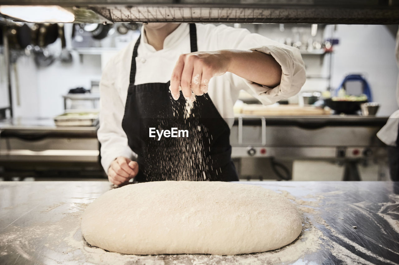 Midsection of female chef student sprinkling flour on dough at commercial kitchen