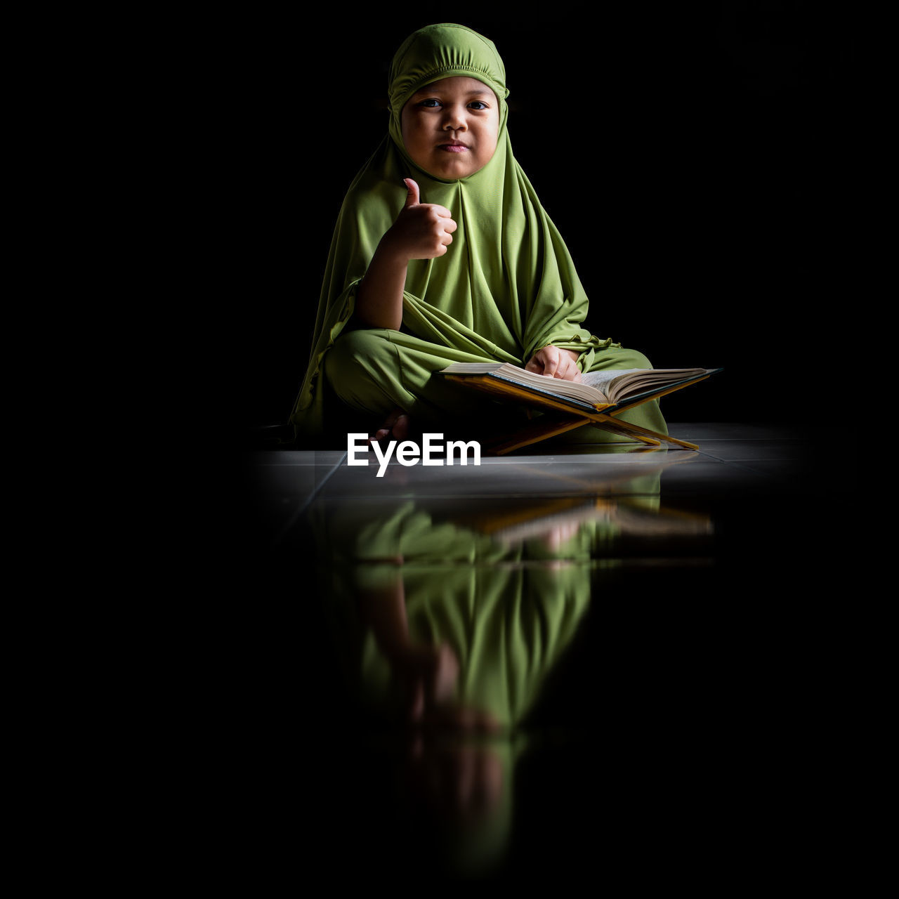 Portrait of girl wearing hijab reading koran while sitting on floor against black background