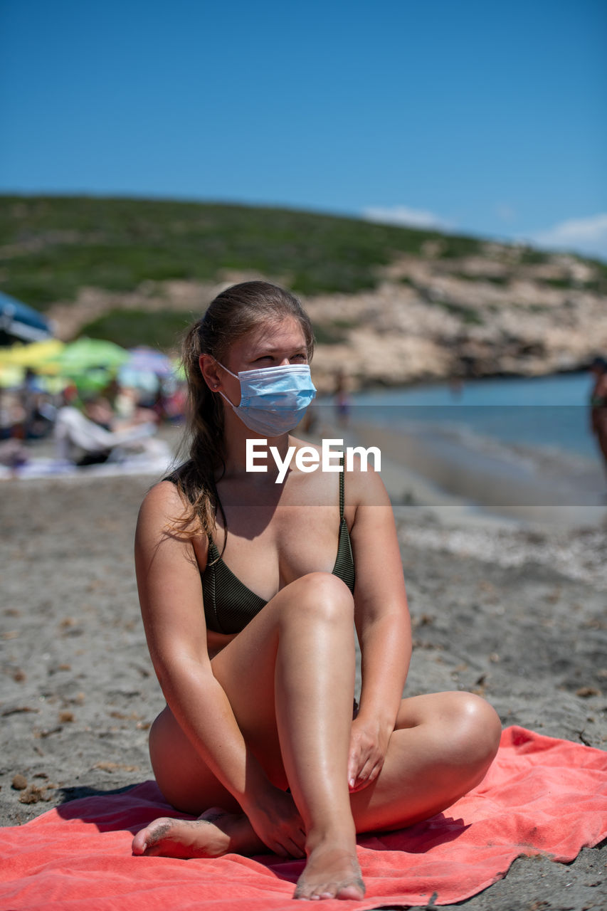 Woman wearing mask in bikini sitting at beach