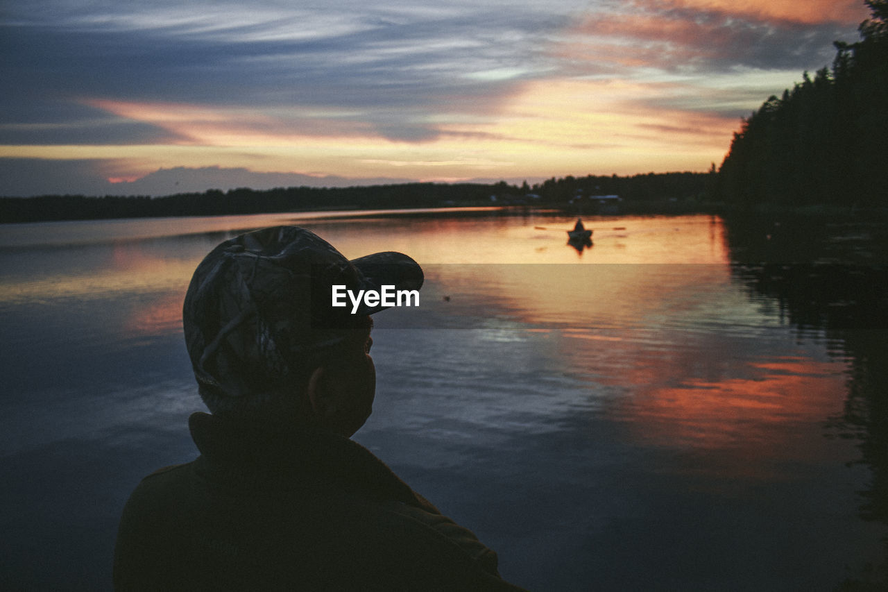 Rear view of man looking away while standing by lake at sunset