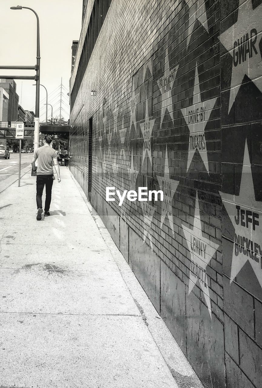 REAR VIEW OF MAN WALKING ON FOOTPATH AGAINST WALL