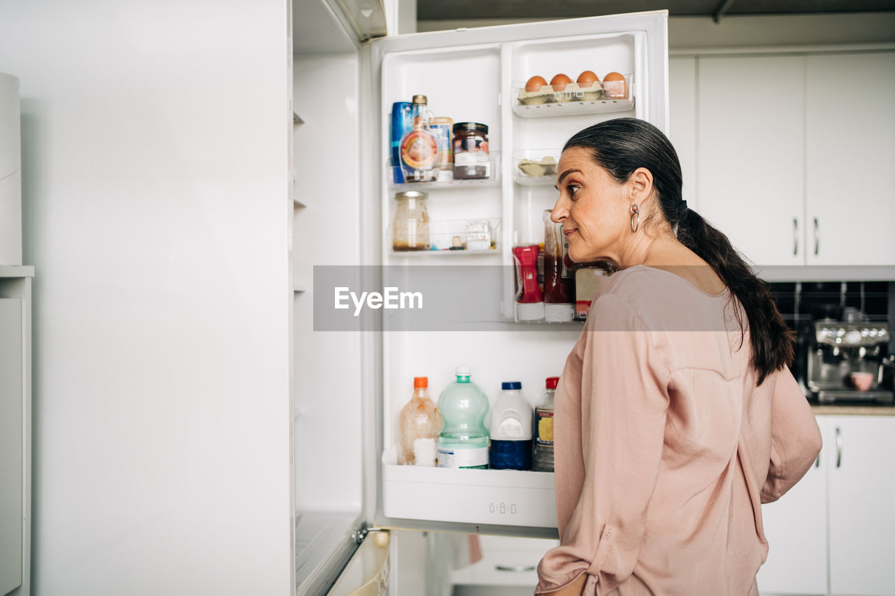 Side view of calm female with ponytail opening door of refrigerator with various products while standing in kitchen with white cupboards