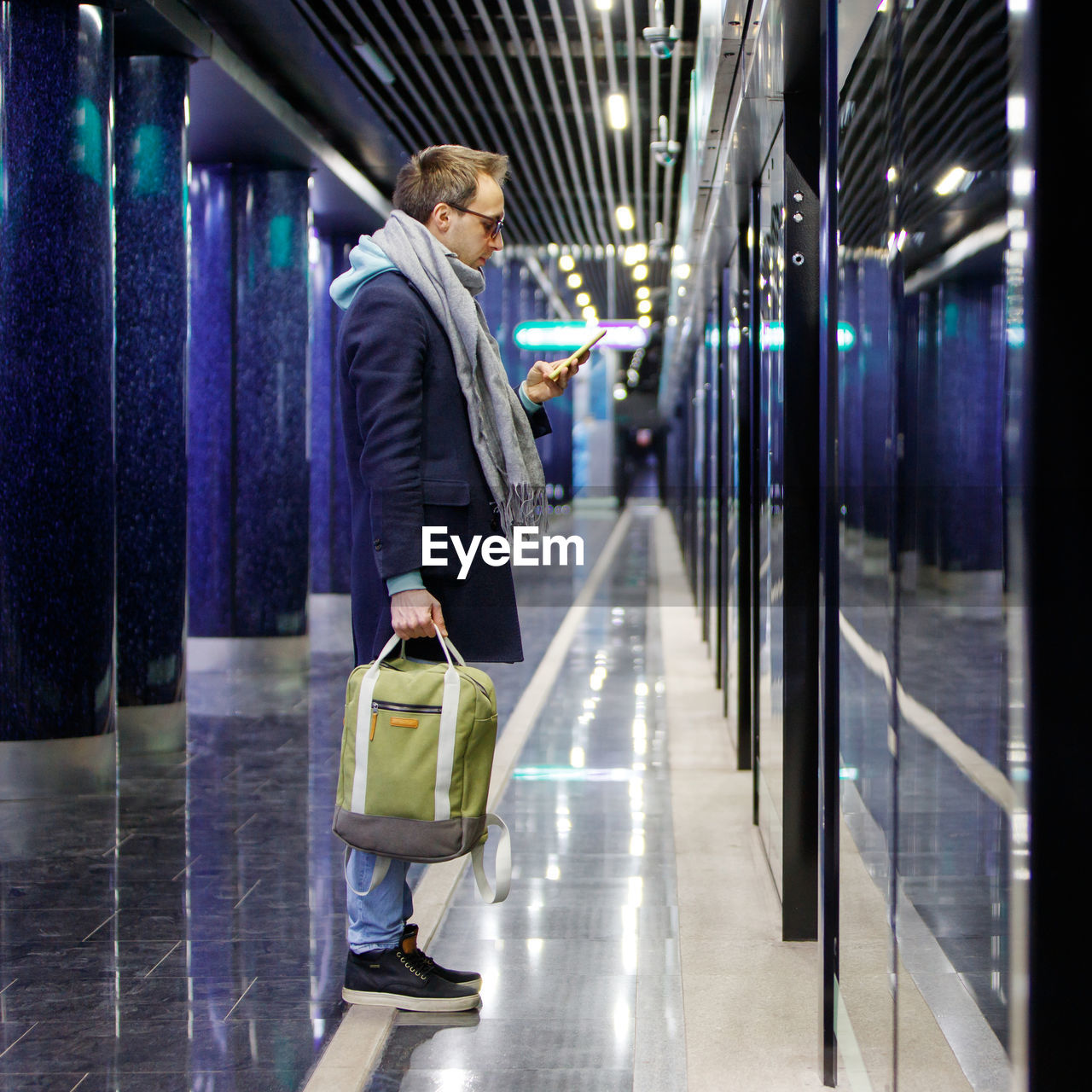 Full length of man using smart phone while standing in illuminated corridor