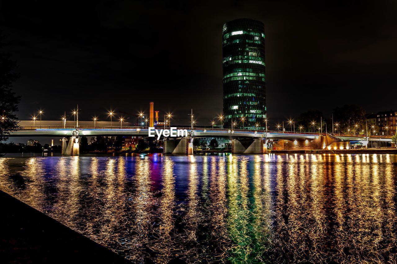 Illuminated bridge over river by buildings against sky at night