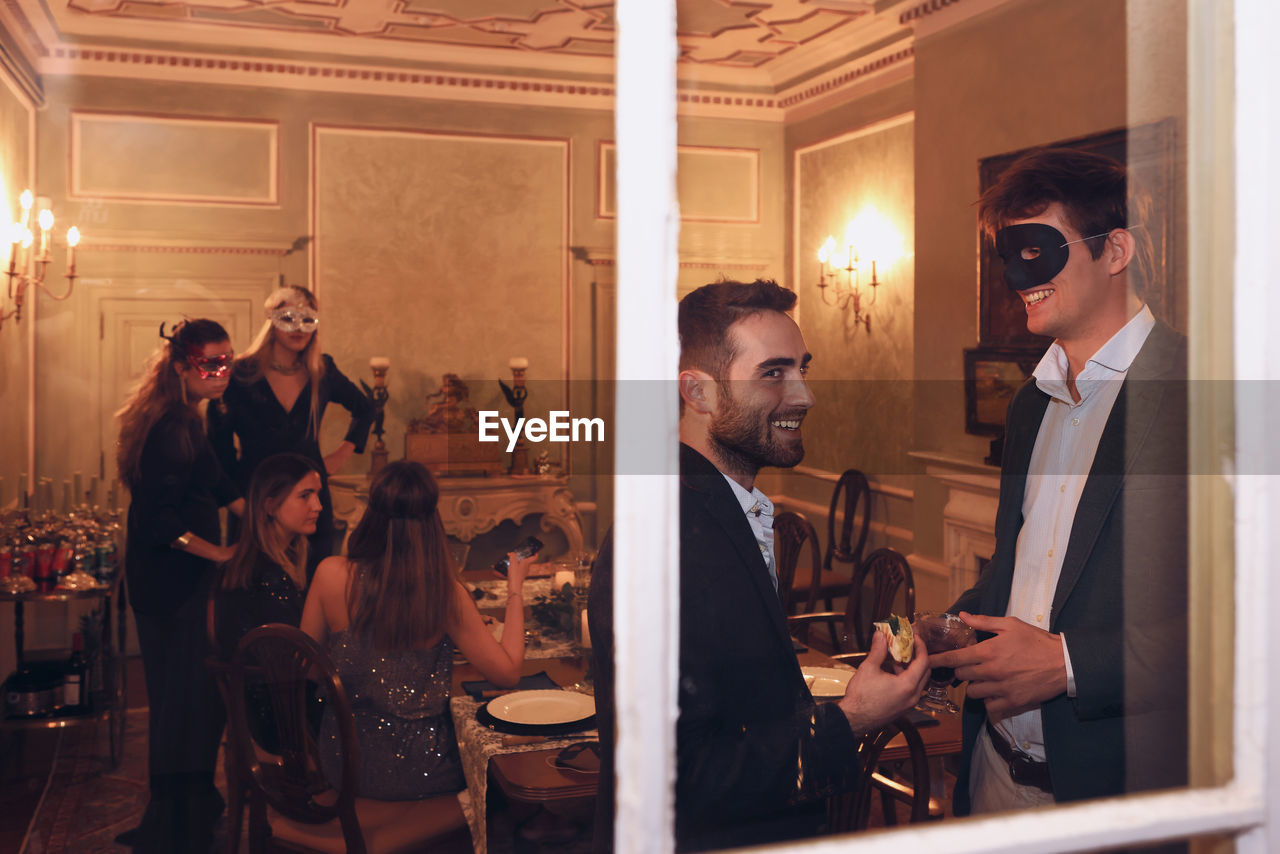 Through window of group of friends in elegant wear and masquerade masks chatting while having dinner together in restaurant