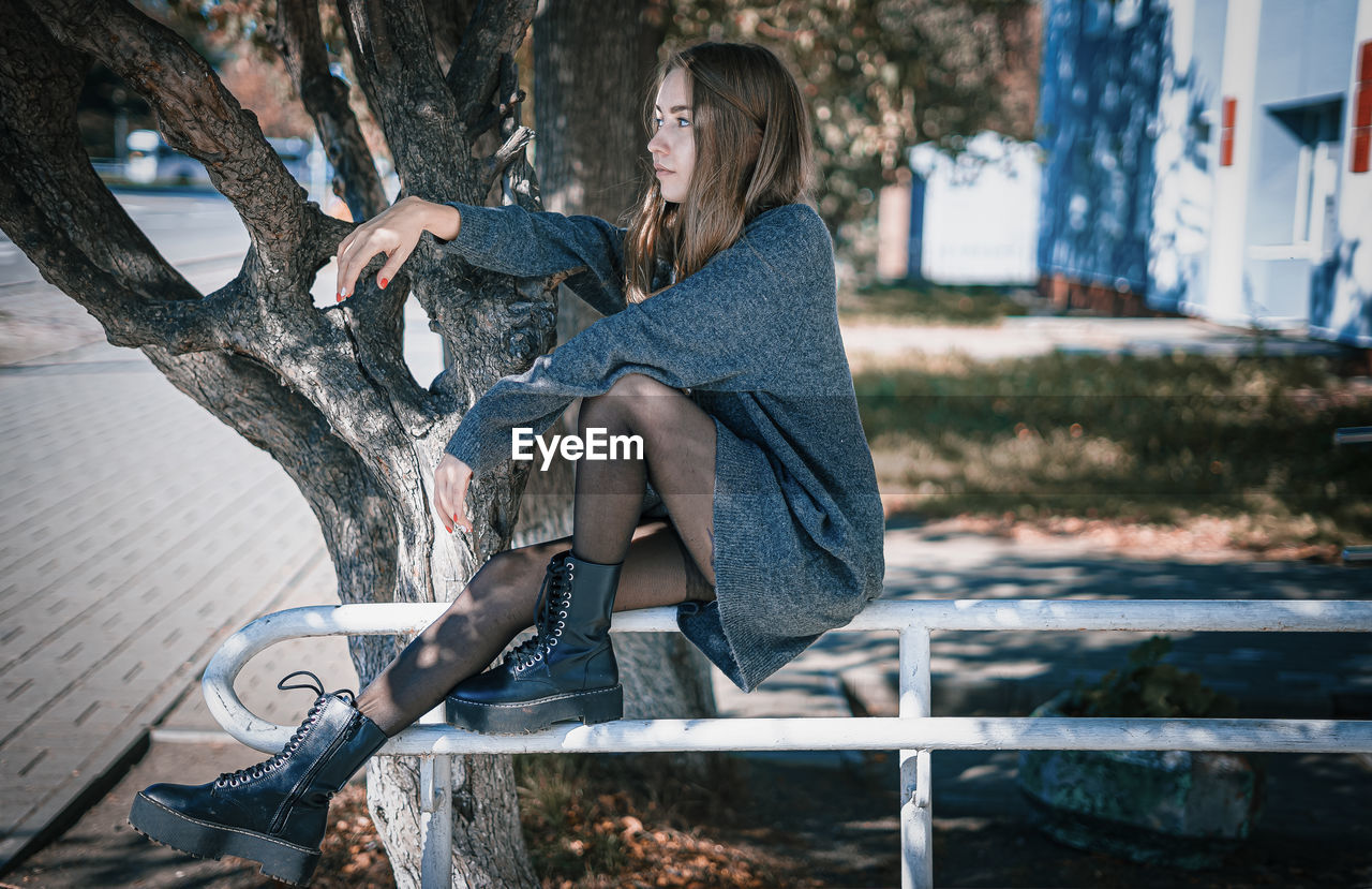 SIDE VIEW OF WOMAN SITTING ON BENCH AGAINST TREE