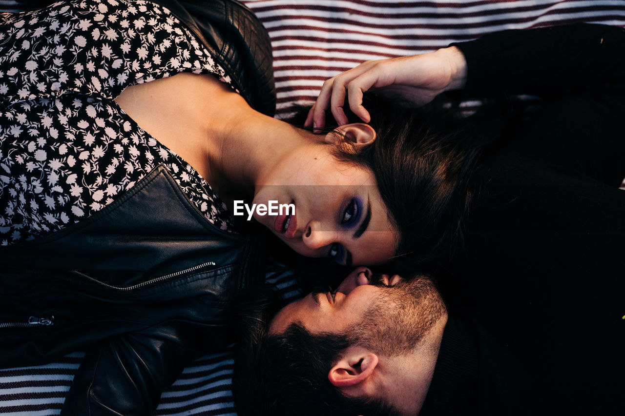 Top view of young ethnic woman with makeup in leather jacket lying near bearded male beloved on textile