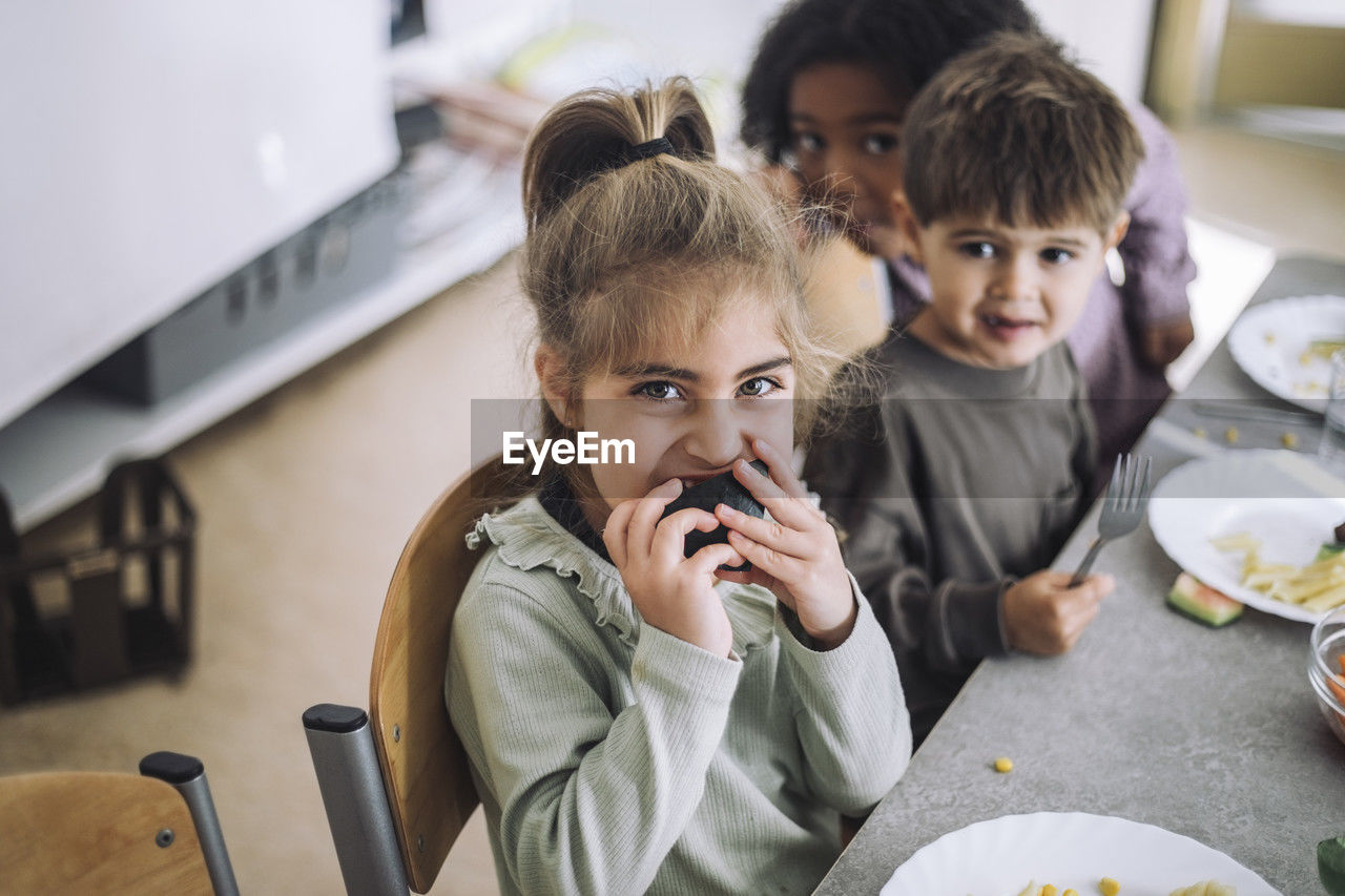 Portrait of girl biting fruit while sitting at dining table with mates in kindergarten