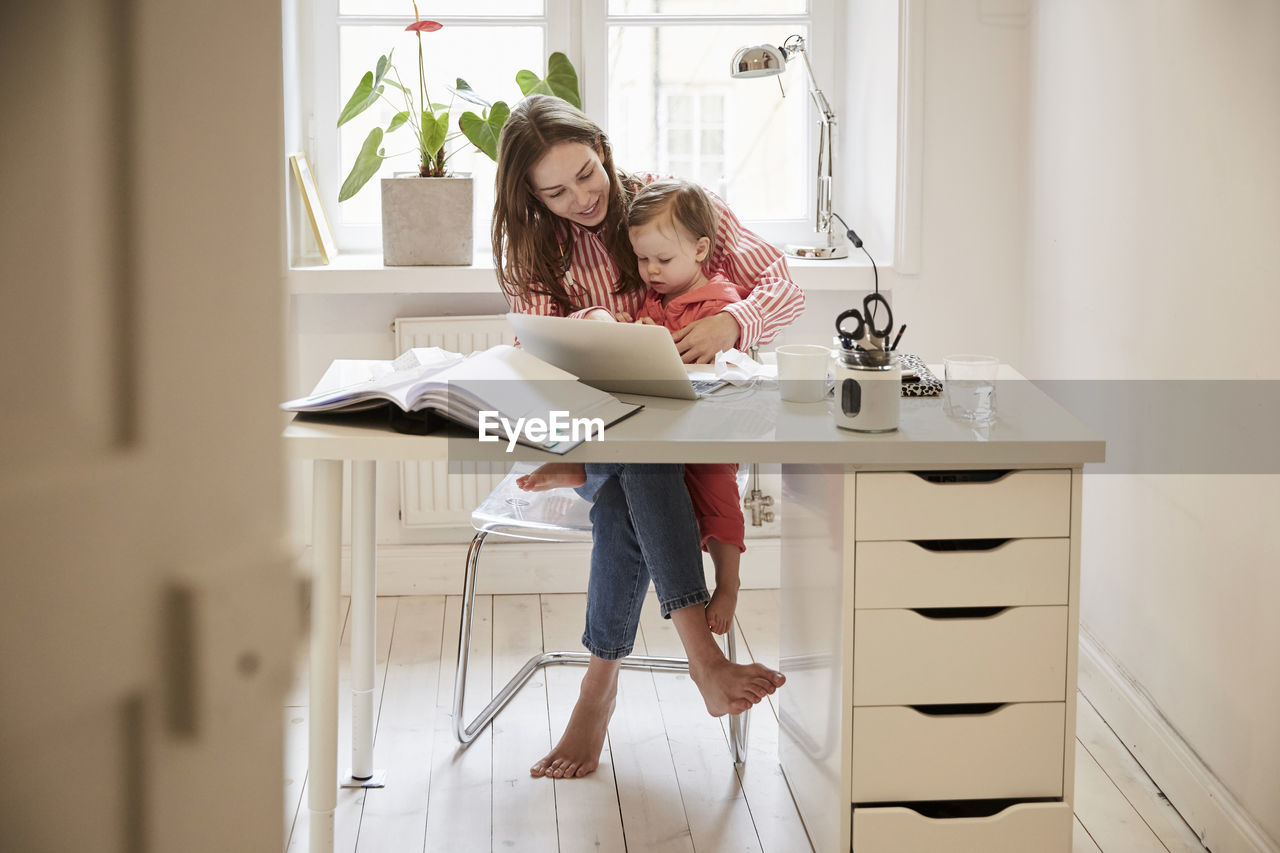 Female accountant sitting with daughter while working on laptop at home