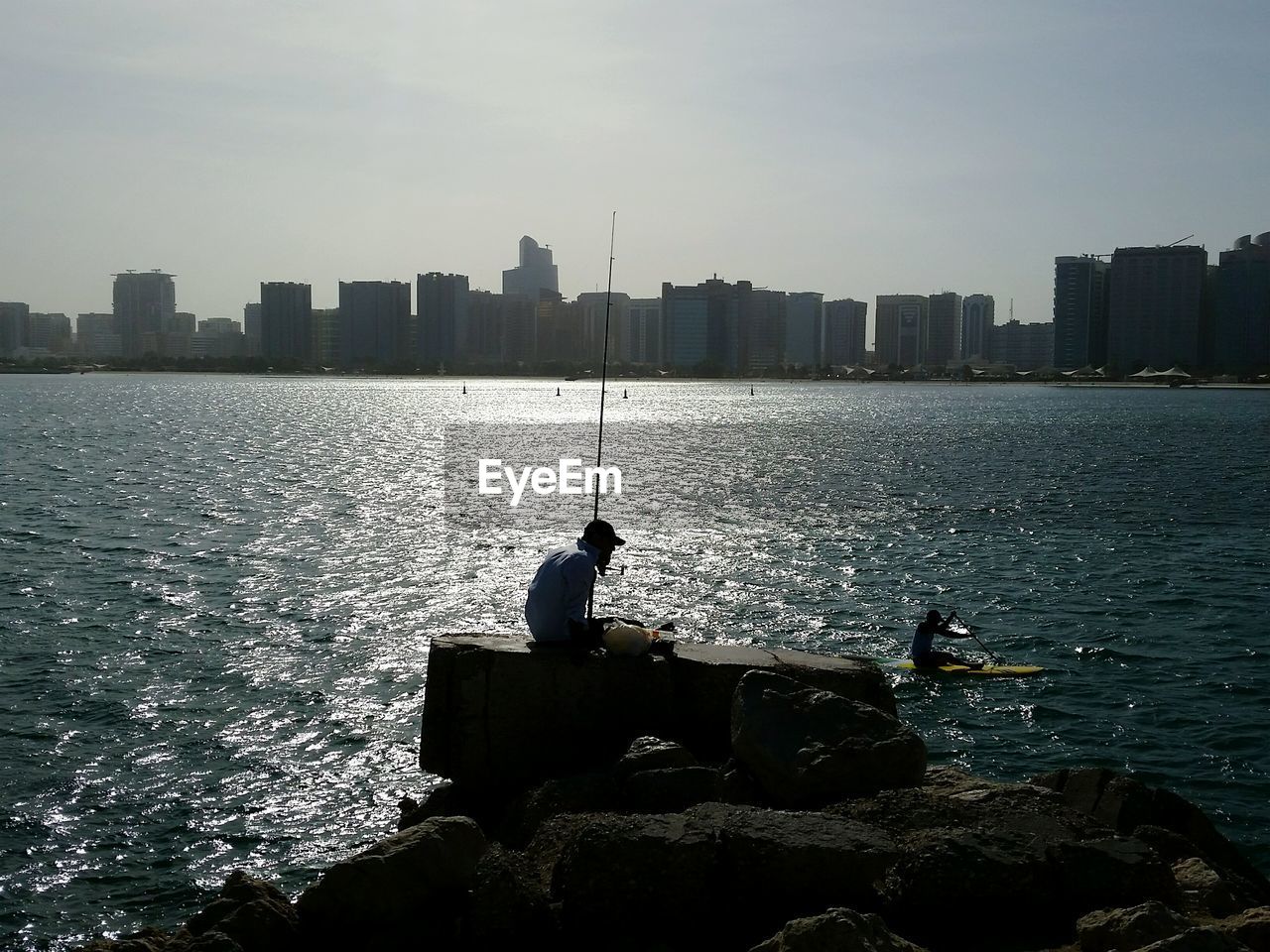 Man on rock while fishing in the sea by city against sky