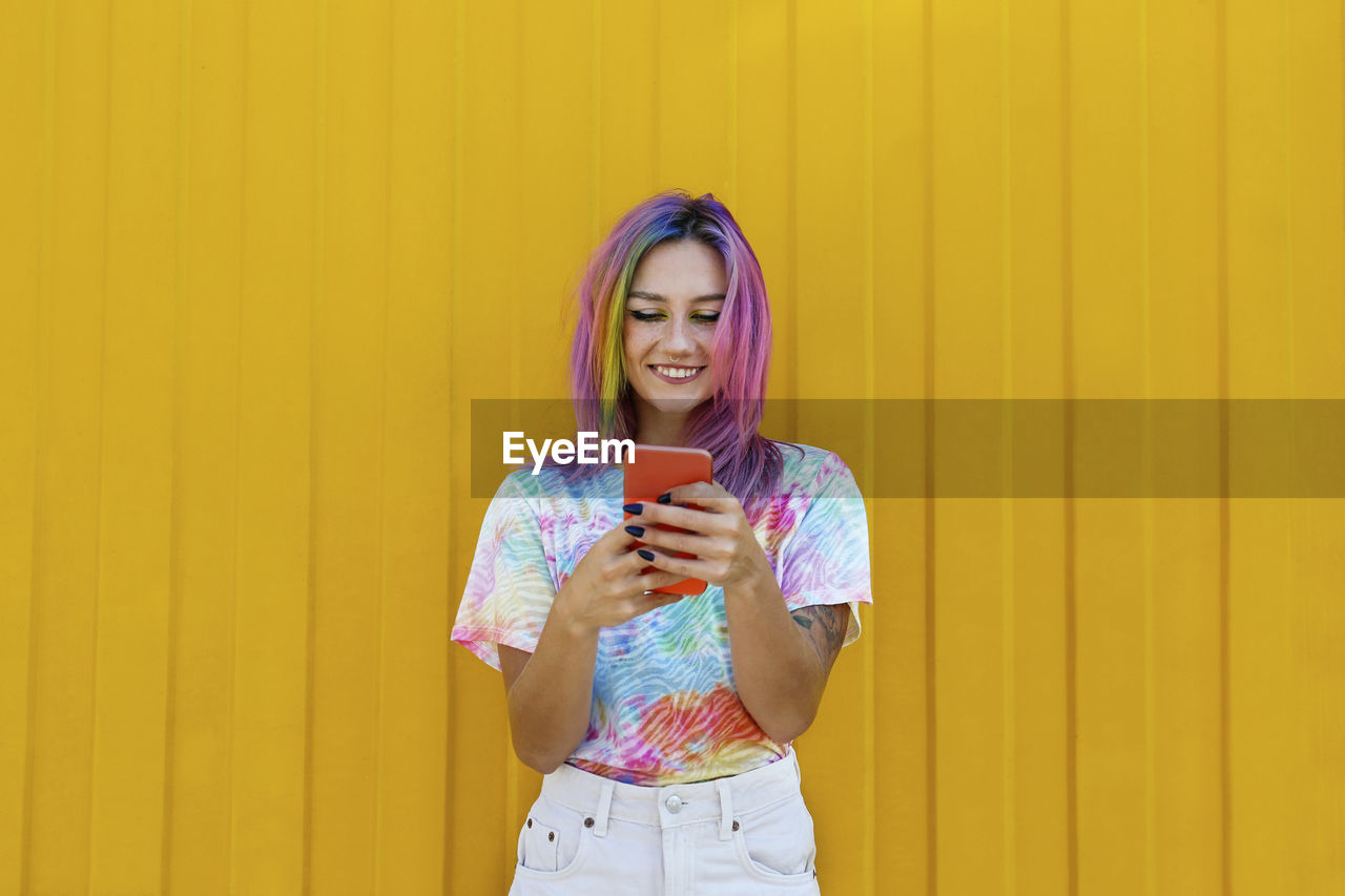 Smiling young woman wearing tie dye t-shirt using smart phone in front of yellow wall