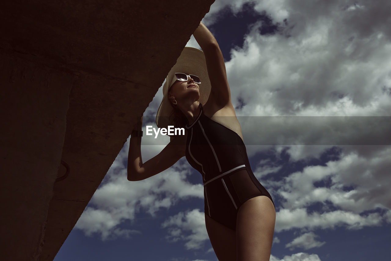 A portrait of a woman in a swimsuit, hat and sunglasses in summer on the riverbank against the sky