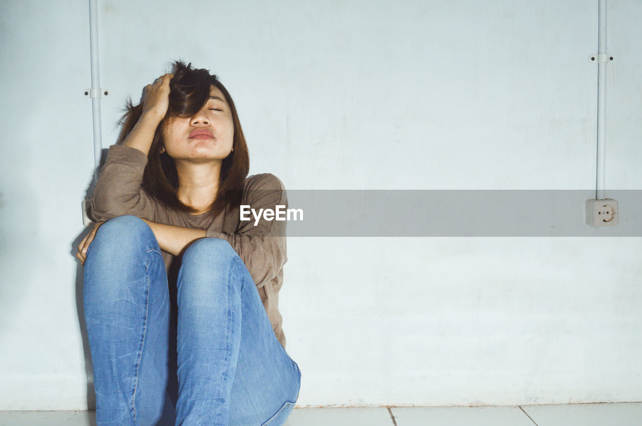 Sad young woman with eyes closed sitting against wall at home