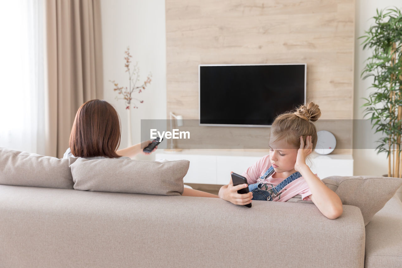 Girl looking at smart phone sitting by mother on sofa at home