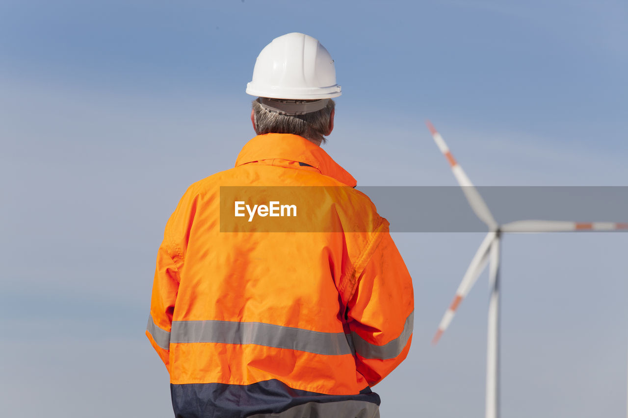 Worker or engineer with hard hat and protecive clothing looking at a windmill of a windfarm