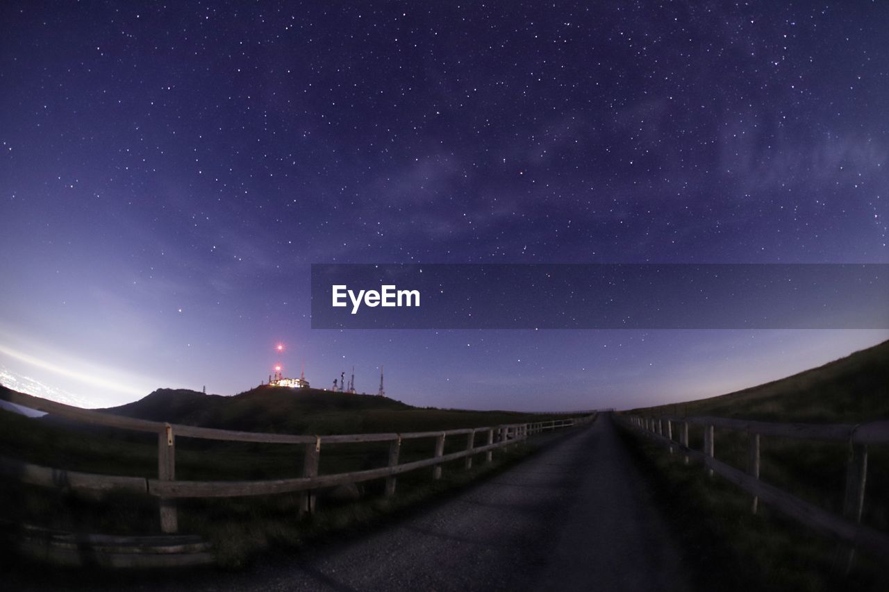 EMPTY ROAD AMIDST ILLUMINATED STAR FIELD AGAINST SKY