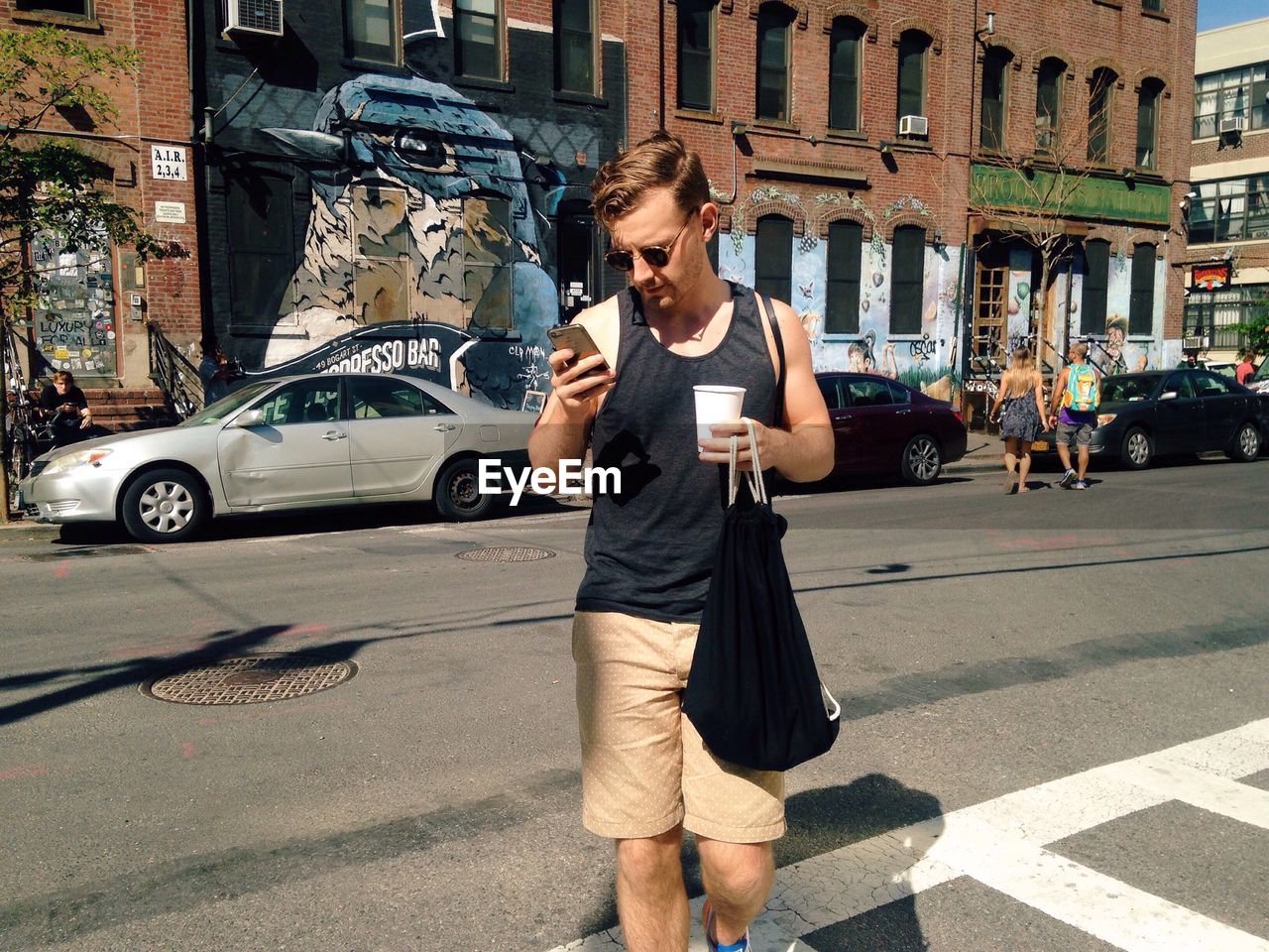 Young man using smart phone while carrying bag and coffee cup on street