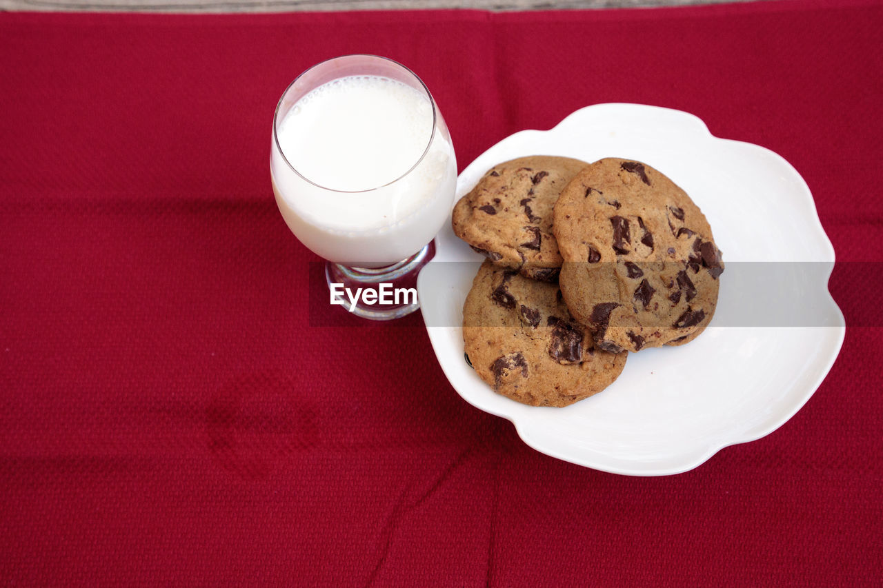 Chocolate chip cookies on a white plate with whole milk in a glass on a red tablecloth