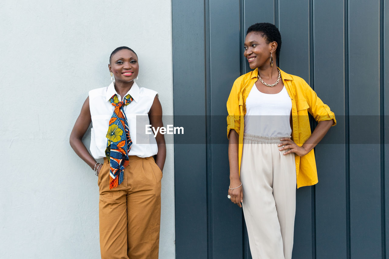 Confident adult african american female friends with short dark hair in stylish outfits smiling and looking at camera while standing against gray wall in city