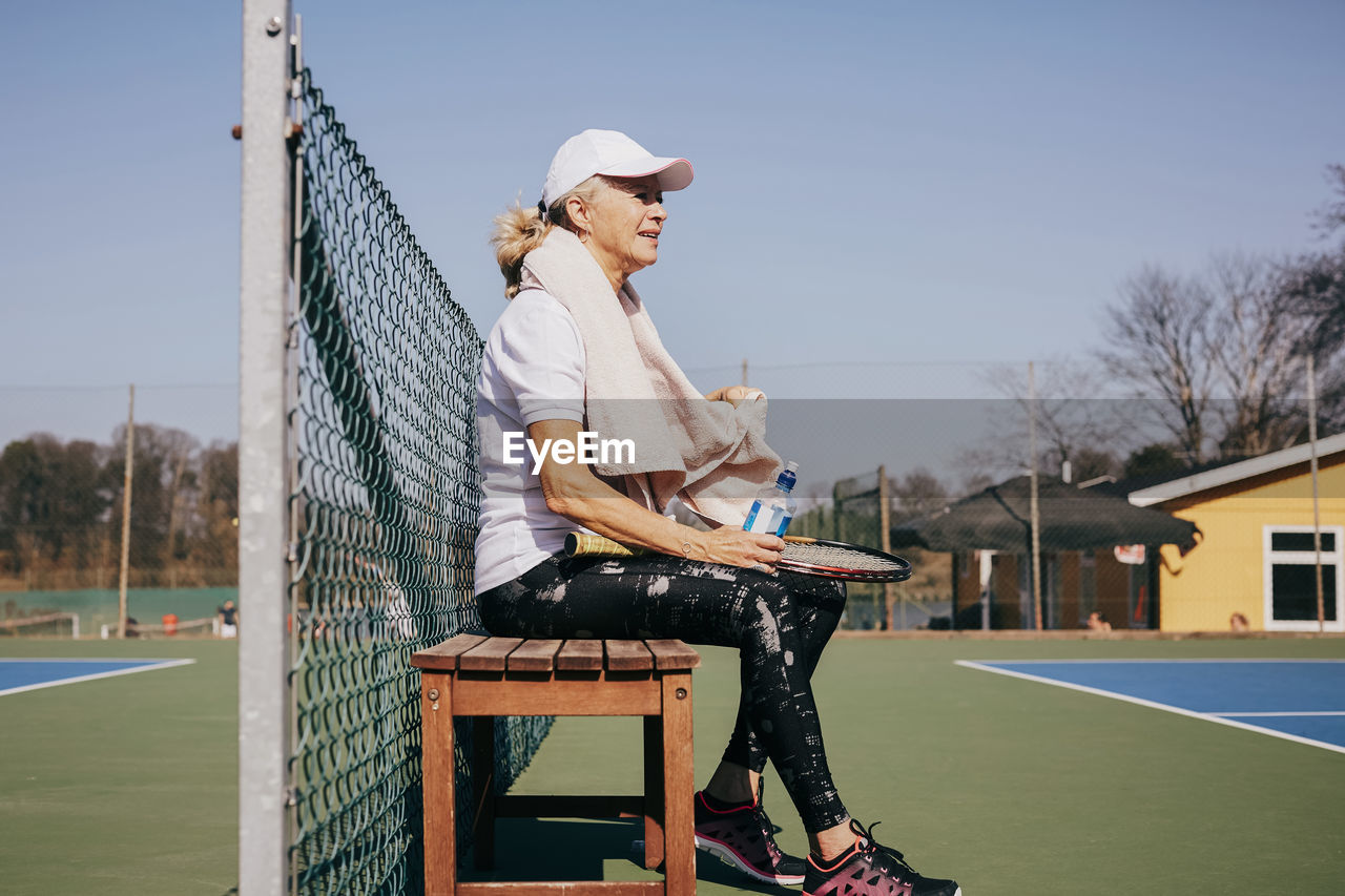 Side view of tired senior woman sitting on bench against sky at tennis court