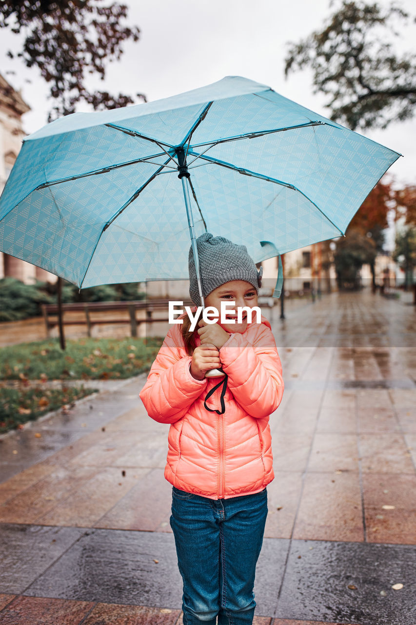 Little smiling happy girl holding big blue umbrella during walk on rainy gloomy autumn day