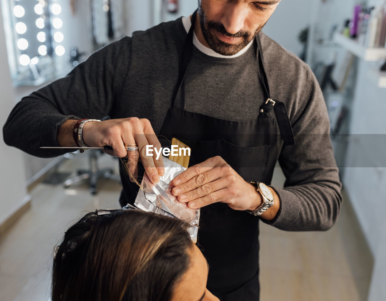 Crop focused ethnic male master combing strands of female customer before dyeing in balayage technique