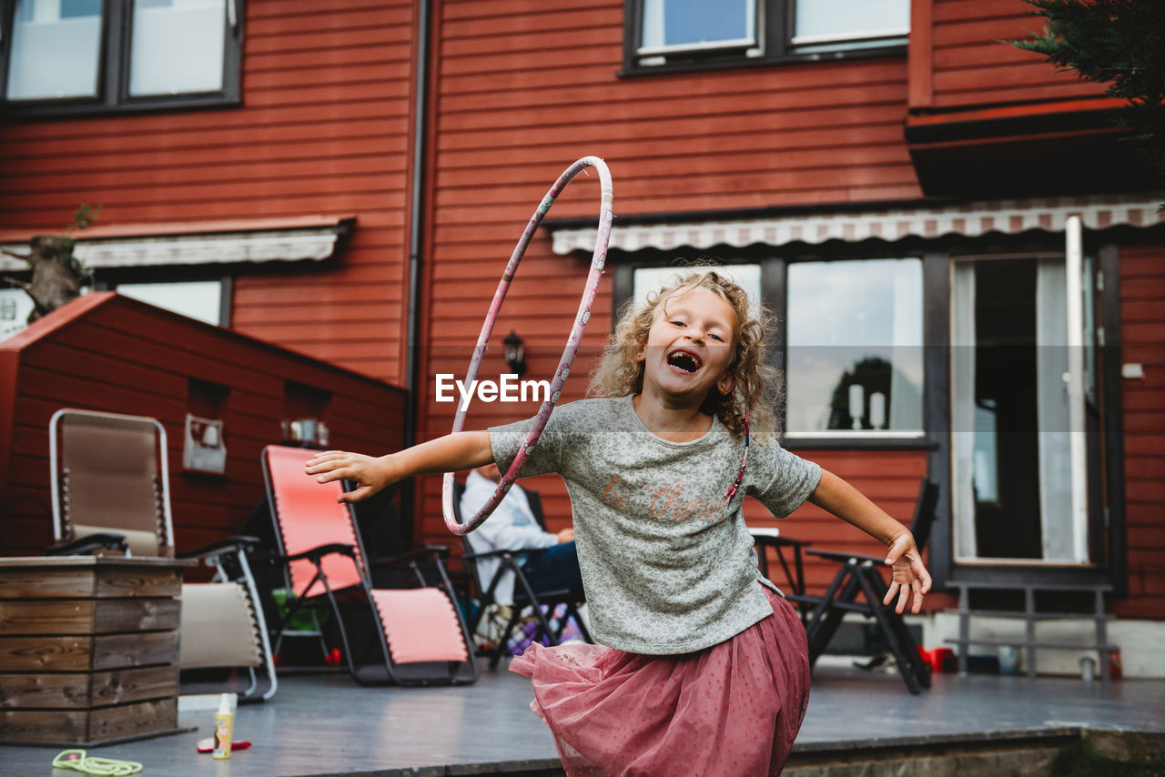 Cute girl having fun with hula hoop in backyard with red wooden house