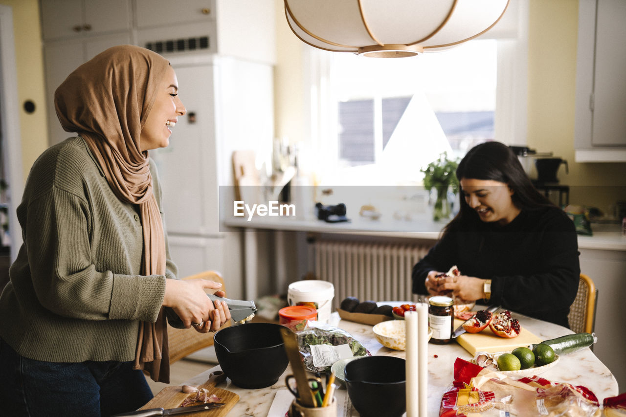 Happy young woman in hijab preparing food with friend sitting at table in kitchen