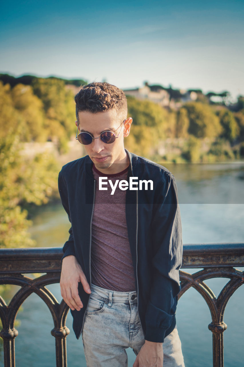 YOUNG MAN WEARING SUNGLASSES STANDING BY RAILING AGAINST RIVER