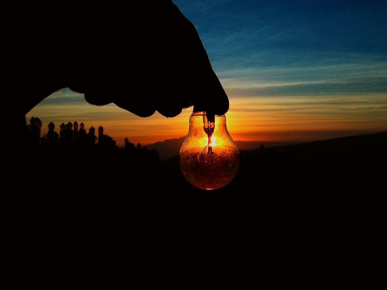 Cropped hand of person holding light bulb against sky during sunset