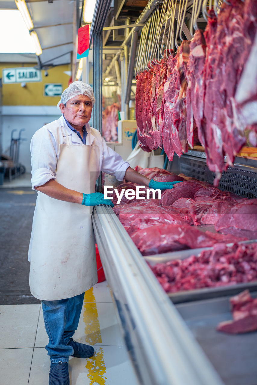 Mature man in uniform touching fresh meat on stall while working in butchery on market