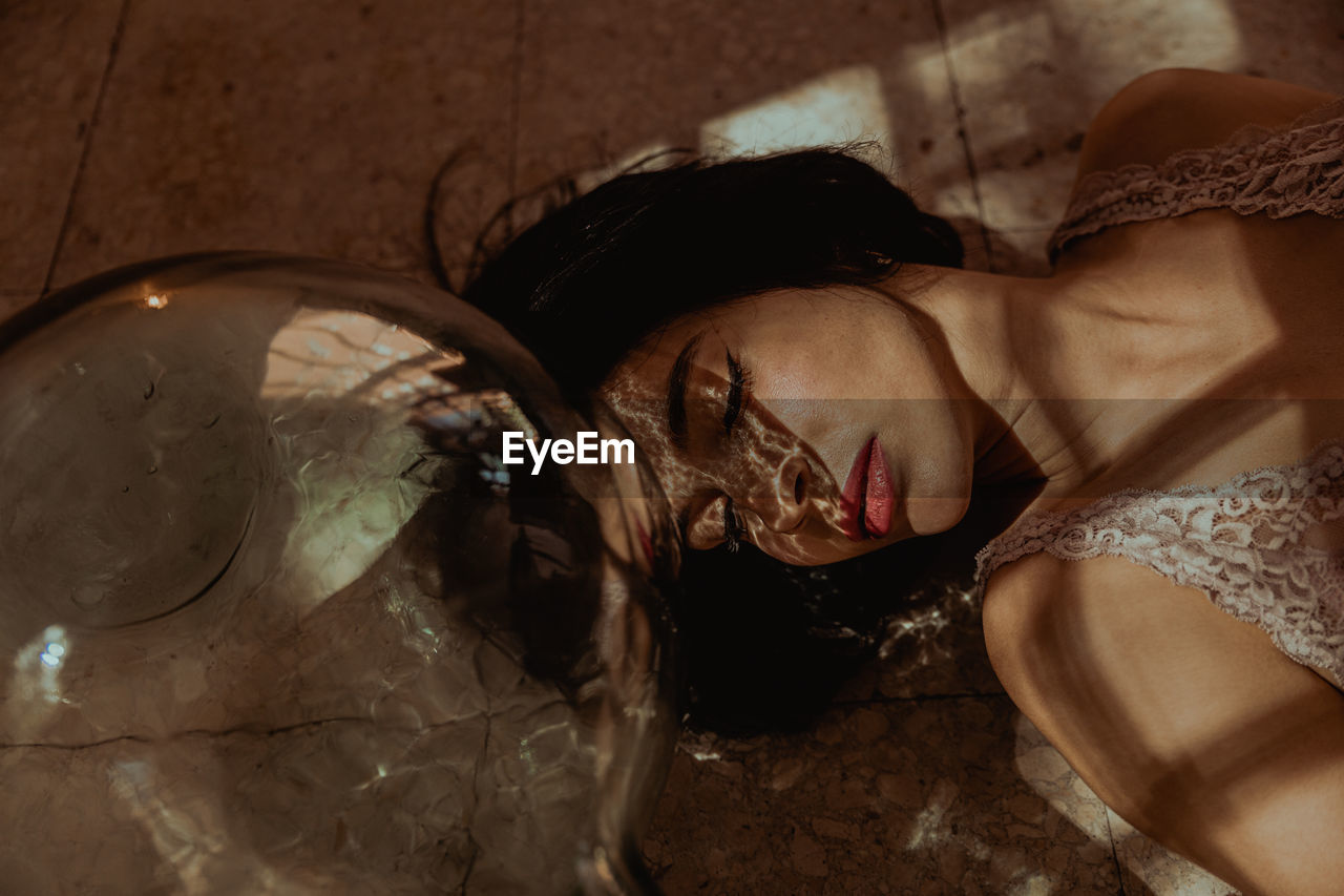 From above of gentle female with closed eyes and in dress lying on floor near glass bottle in room with sunlight