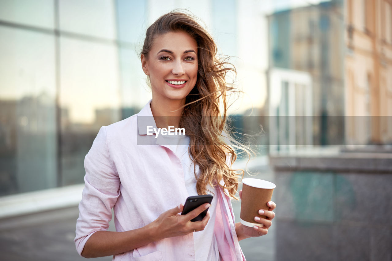 Portrait of smiling woman holding smart phone and coffee while standing in city