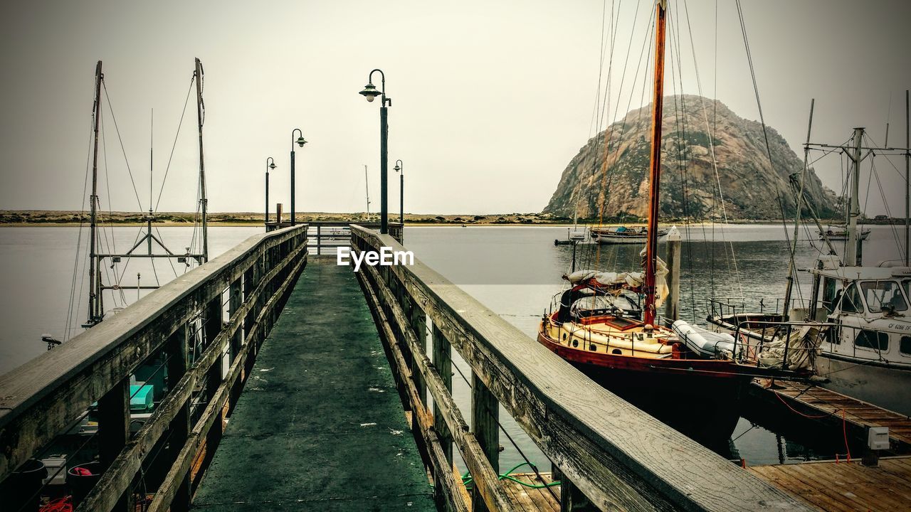 Walkway by moored sailboats against clear sky