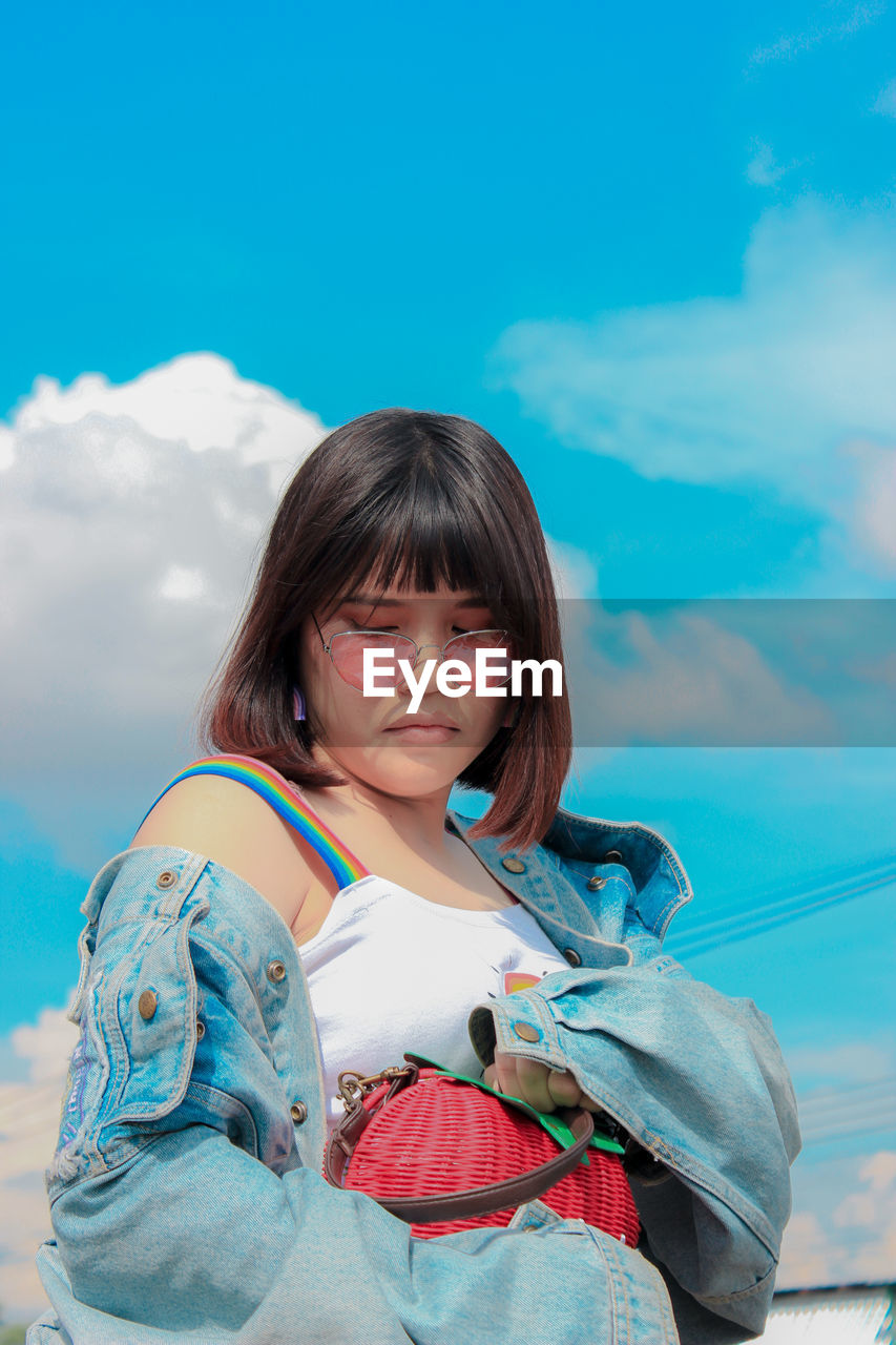 Low angle view of young woman holding purse while standing against blue sky