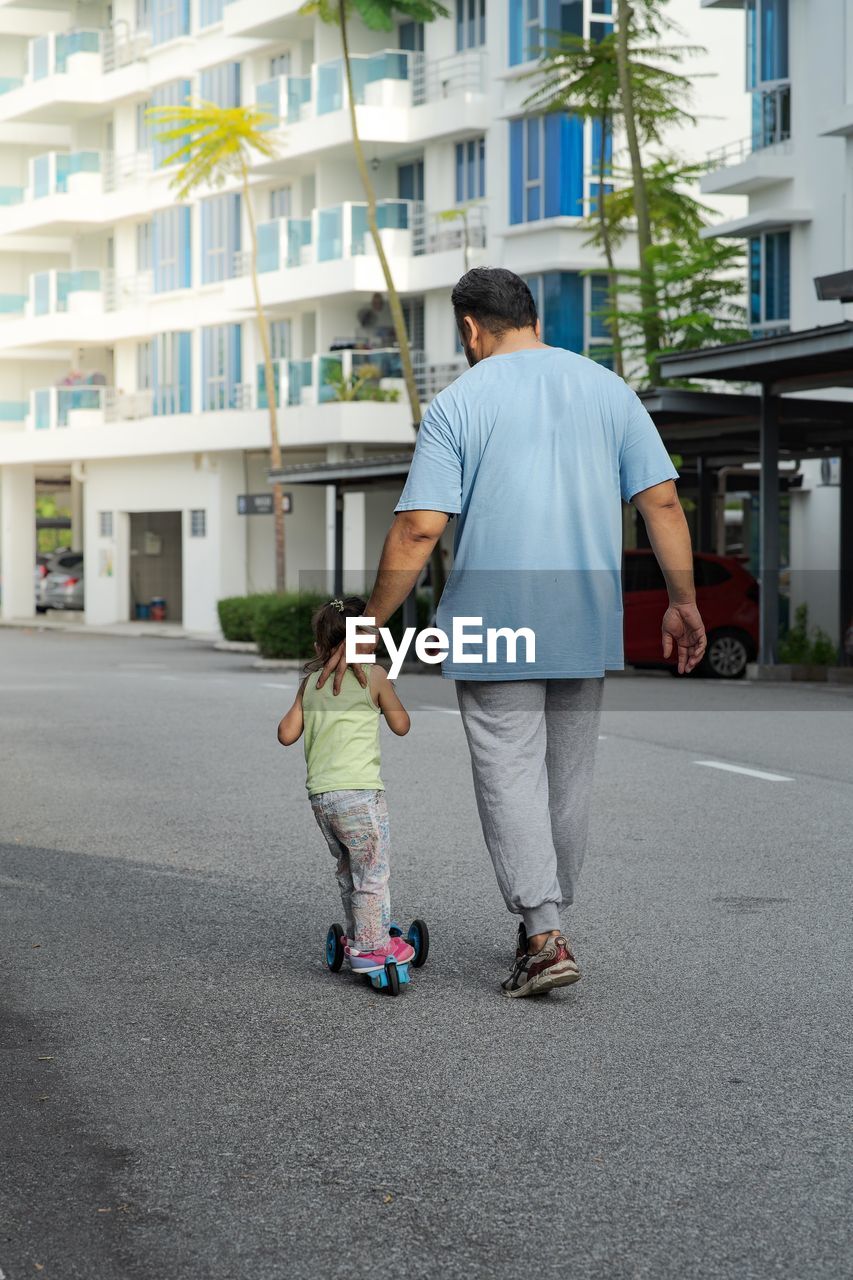 Father walking with girl riding push scooter on road in city