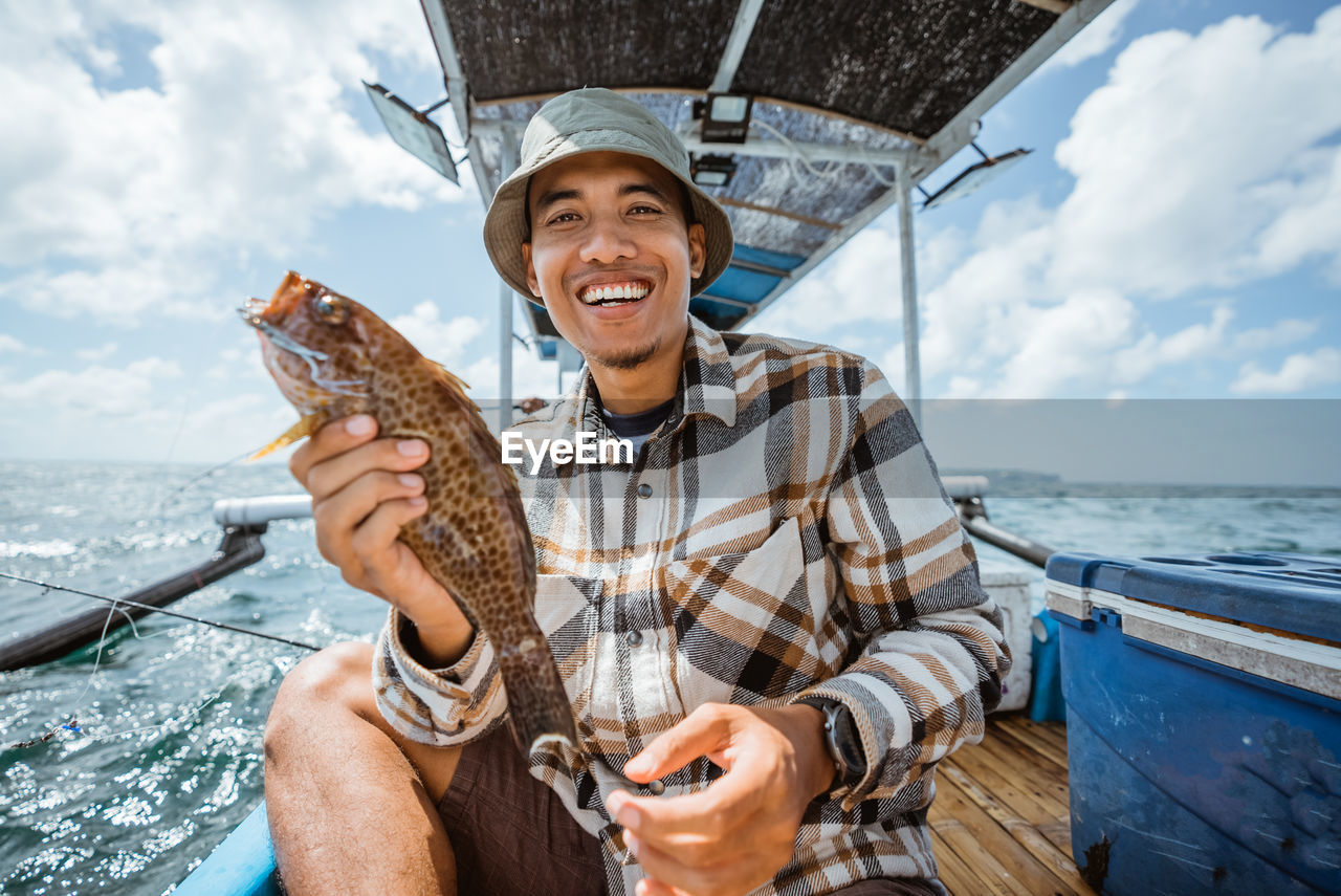 water, sea, adult, smiling, nautical vessel, happiness, transportation, emotion, one person, men, fish, nature, vacation, cloud, mode of transportation, trip, sky, travel, holiday, ship, portrait, cheerful, leisure activity, holding, front view, person, vehicle, big-game fishing, teeth, day, enjoyment, smile, looking at camera, fishing, fun, animal, outdoors, sailor, boat, sailing, hat, mature adult, travel destinations, senior adult, sitting, food and drink, summer, positive emotion, adventure, boat deck, clothing, sailboat, casual clothing, relaxation, waist up