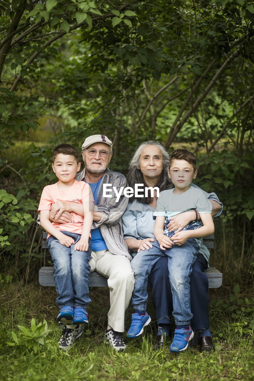 Portrait of grandparents sitting with twin grandsons on bench against plants in back yard