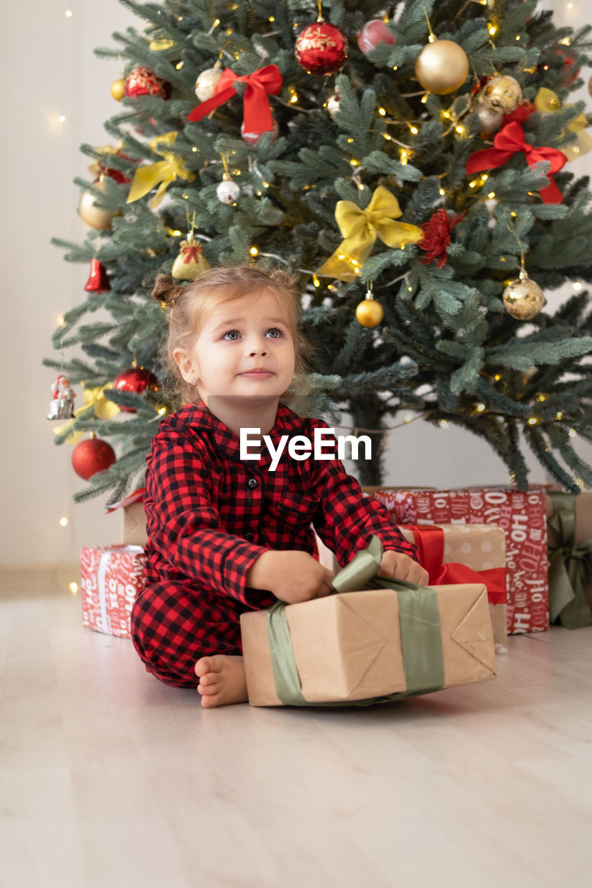 Cute little child girl in red pajama with gift boxes sitting under christmas tree at home