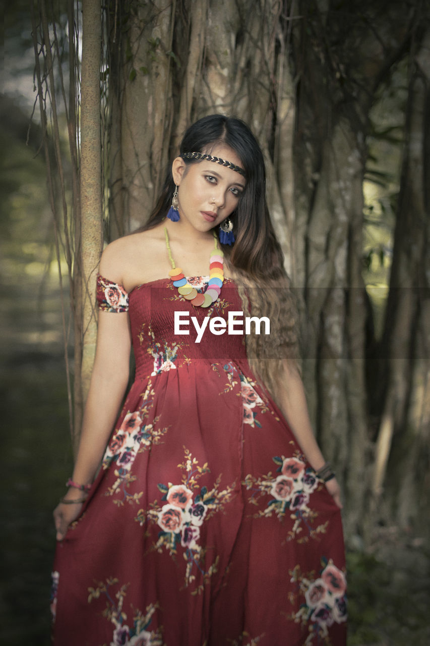 Portrait of woman wearing dress standing against tree trunk in forest