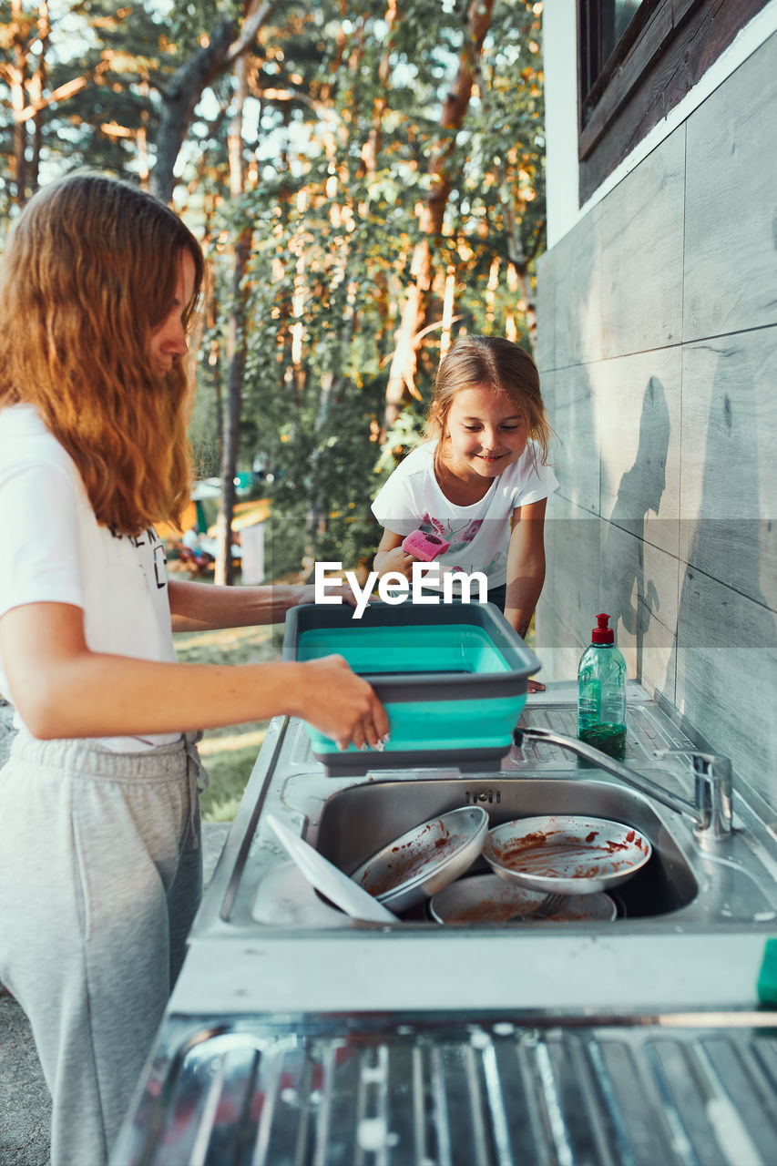 Teenager girl washing up the dishes pots and plates with help her younger sister