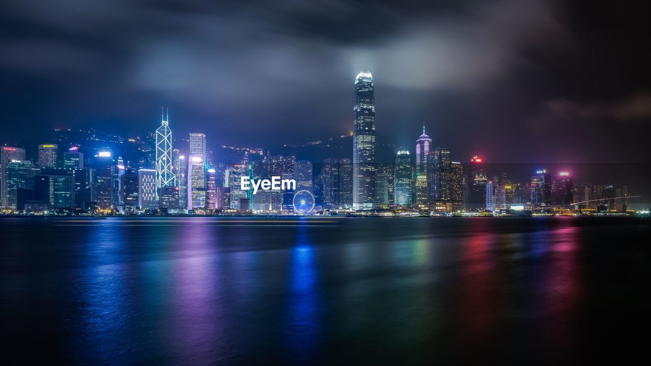 Modern illuminated buildings in front of sea against sky at harbor