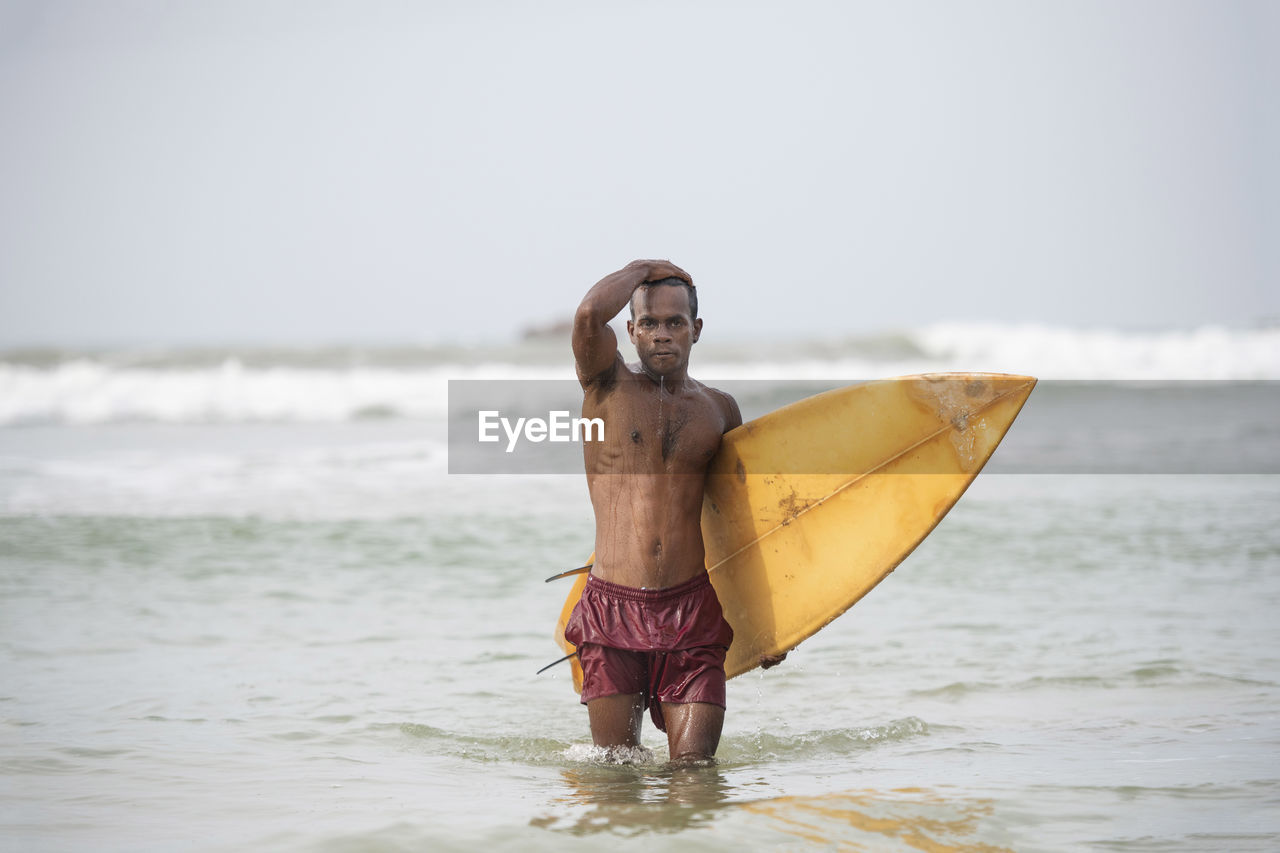 Portrait of shirtless man with surfboard wading in sea against sky