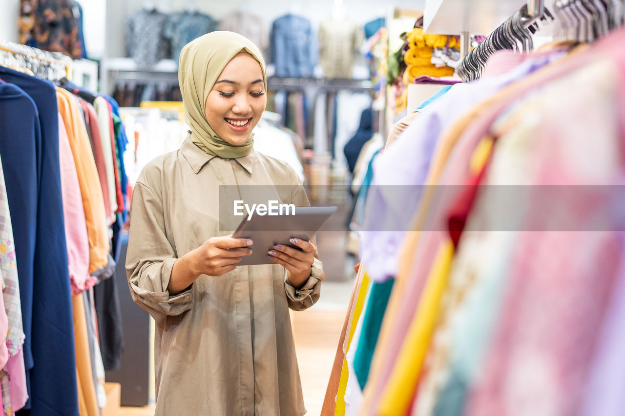 Smiling woman in hijab using digital tablet in clothing store