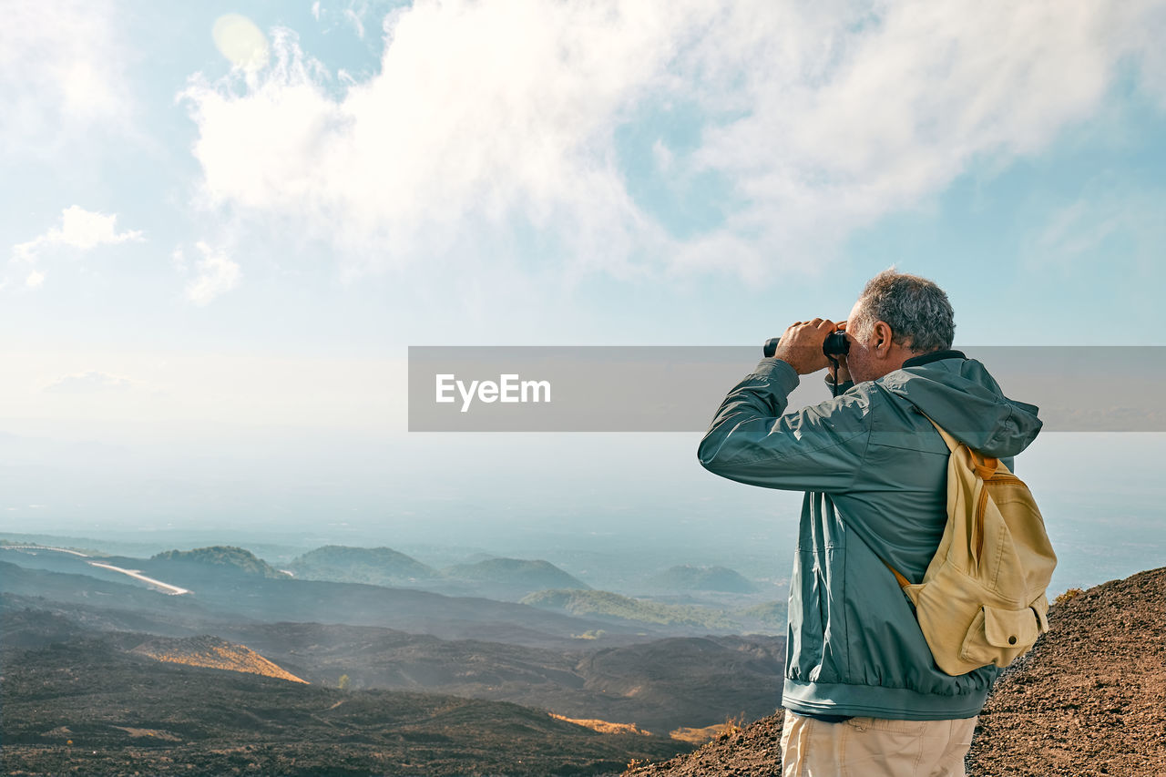 Rear view of man looking through binoculars at panoramic view of summits volcano etna