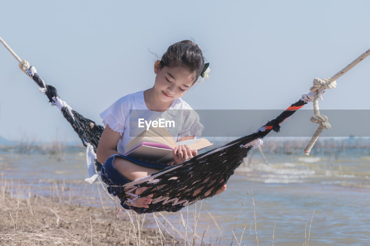 Girl reading book while sitting on hammock against sky