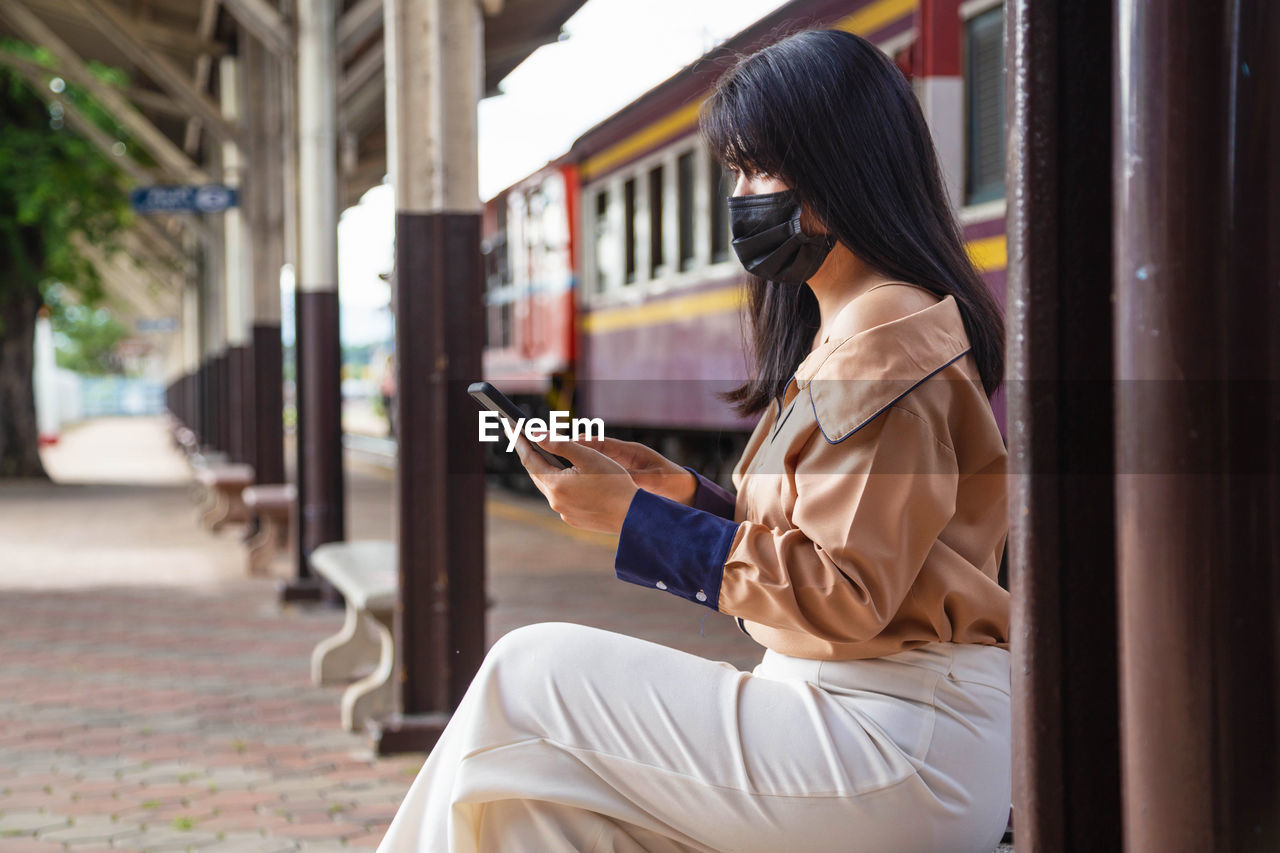 Side view of woman wearing flu mask using mobile phone at railroad station
